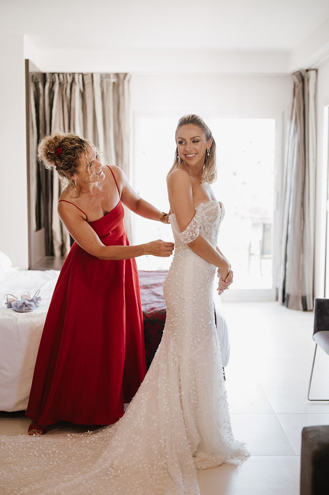 Mother in red gown helping bride fasten her beaded white wedding dress in a bright hotel room.