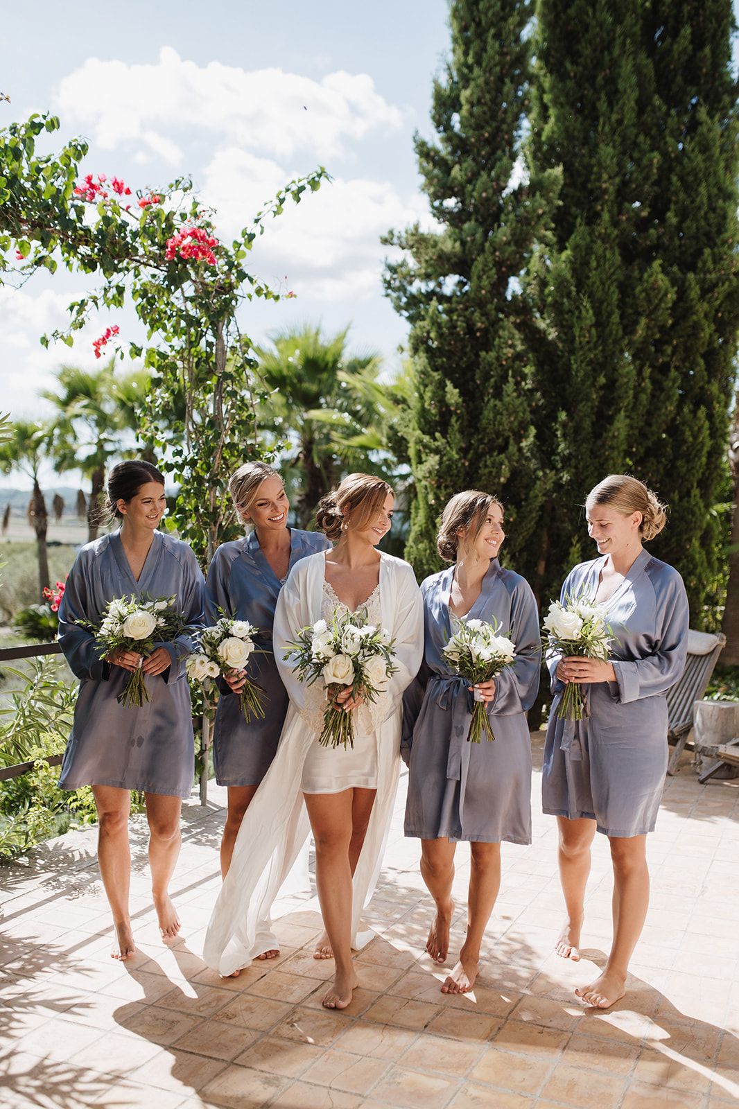 Bride and bridesmaids in grey robes holding white bouquets walking outdoors on a sunny wedding day.