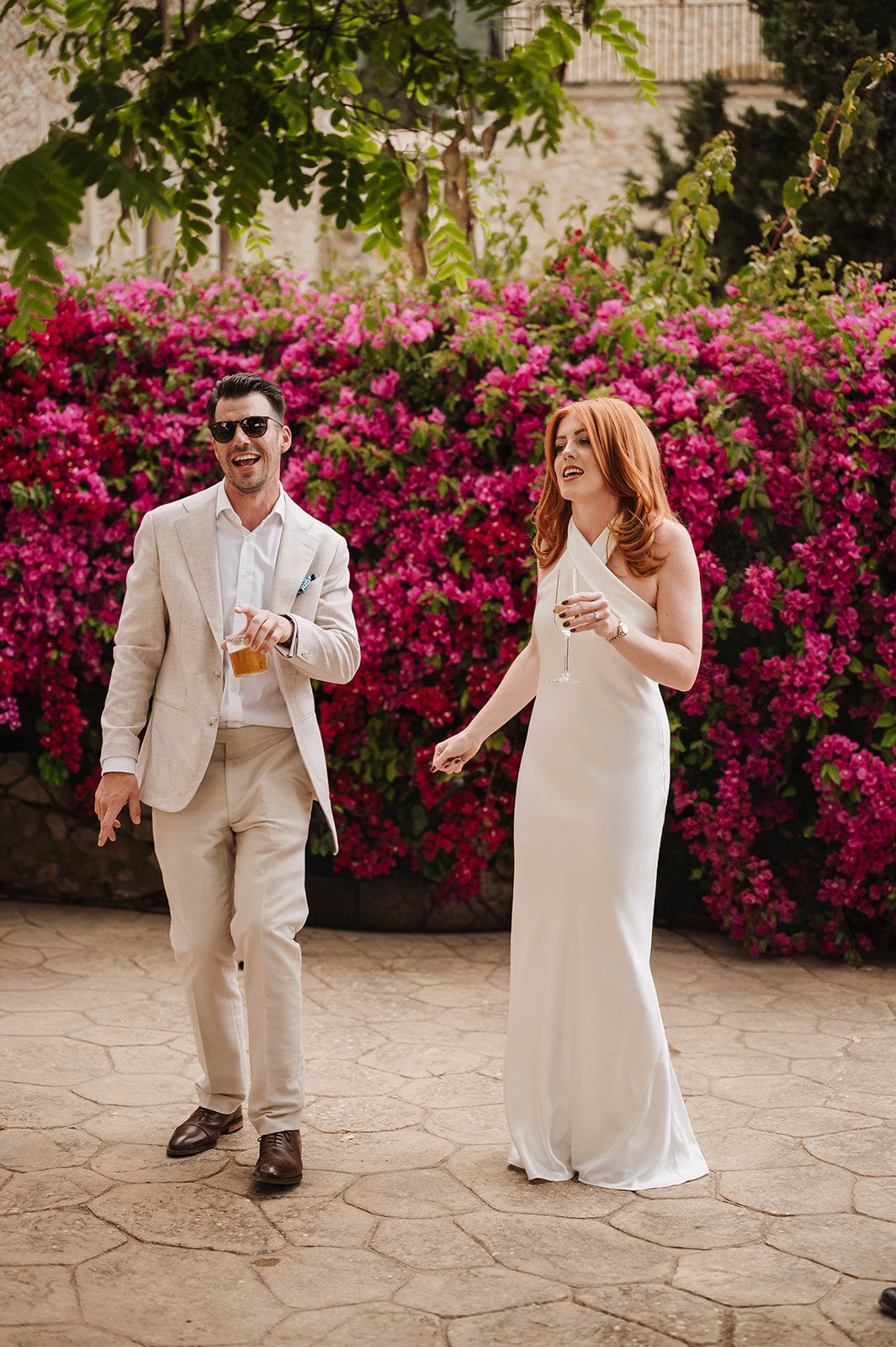 Bride in white gown and groom in beige suit walking and laughing before vibrant pink bougainvillea flowers.
