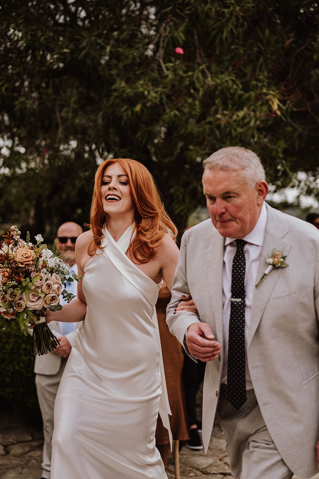 Joyful red-haired bride in sleek white halter dress laughs while walking with father at wedding.