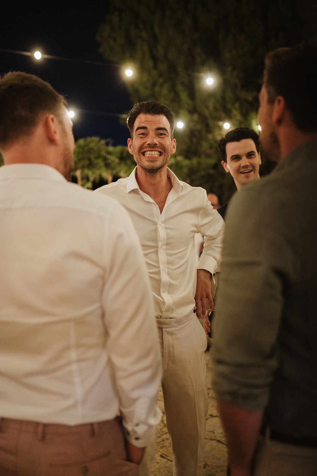 Smiling groom in white shirt and beige trousers socializing with guests under string lights at night.