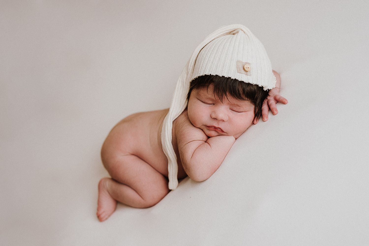 Sleeping newborn baby wearing a cream knit bonnet hat, curled up peacefully on a soft white blanket.