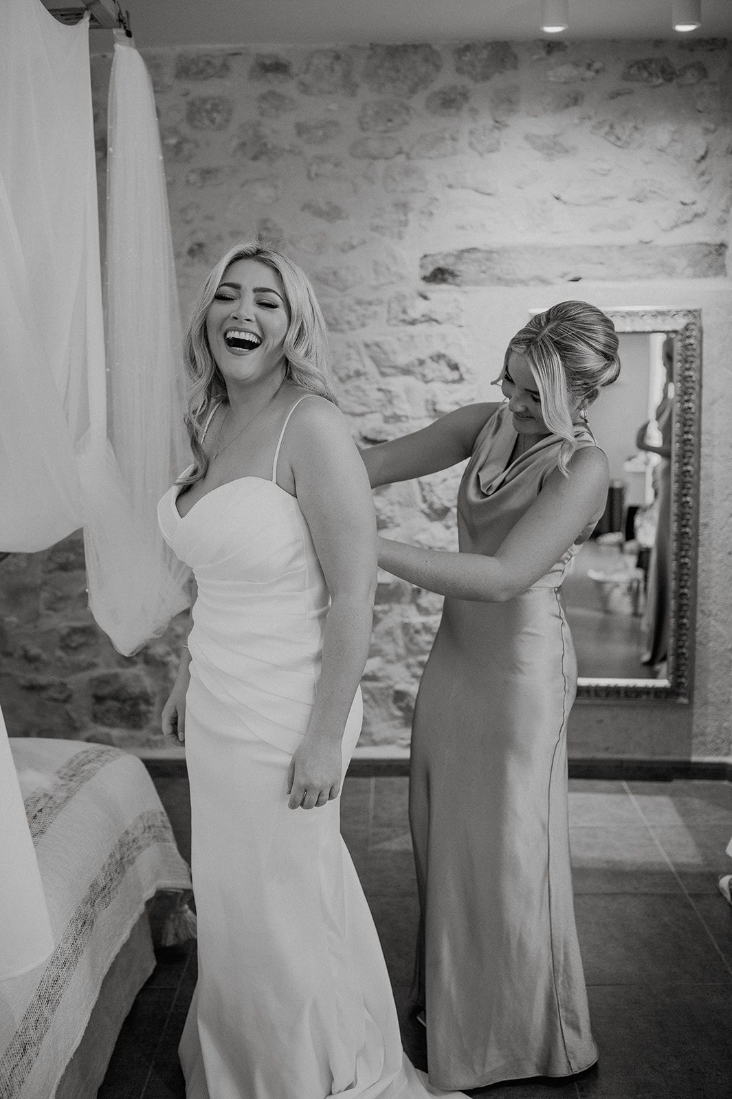 Bride laughing joyfully as bridesmaid helps zip up elegant white wedding gown in rustic stone venue.
