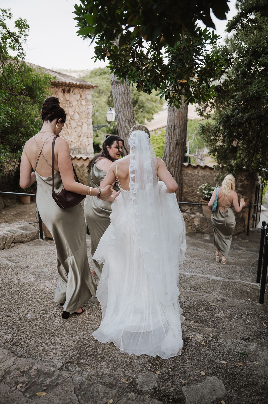 Bridesmaids in sage satin gowns assist bride in white wedding dress and veil walking up stone steps outdoors.