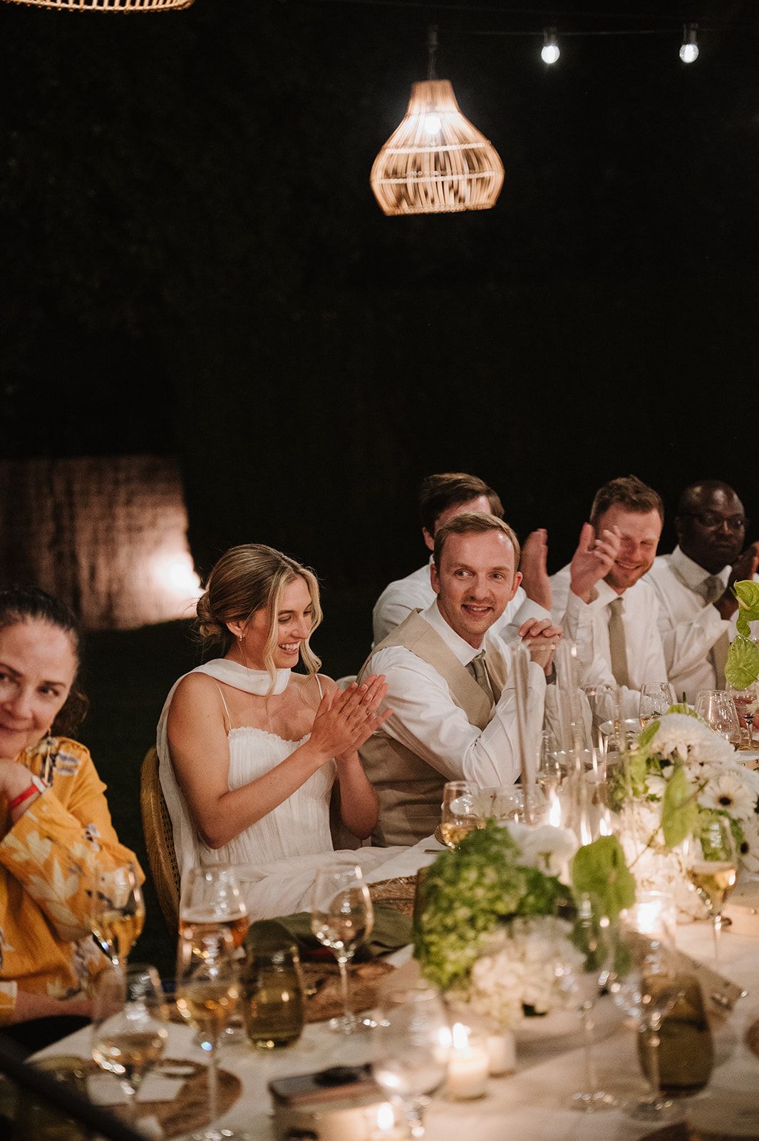 Bride and groom clapping joyfully at candlelit wedding reception table with green floral centerpieces.