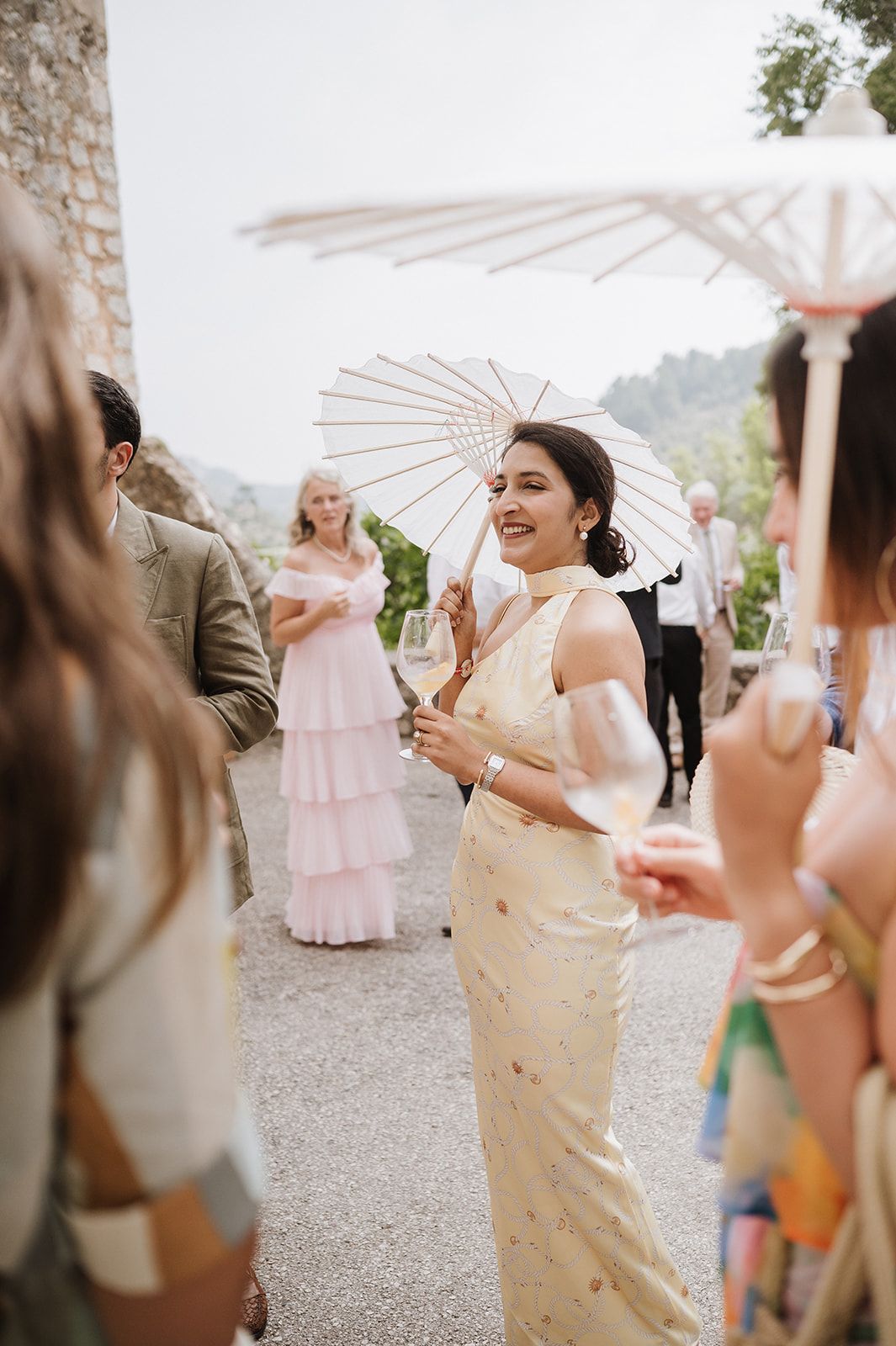 Wedding guest in gold saree holds white parasol while socializing at outdoor reception.