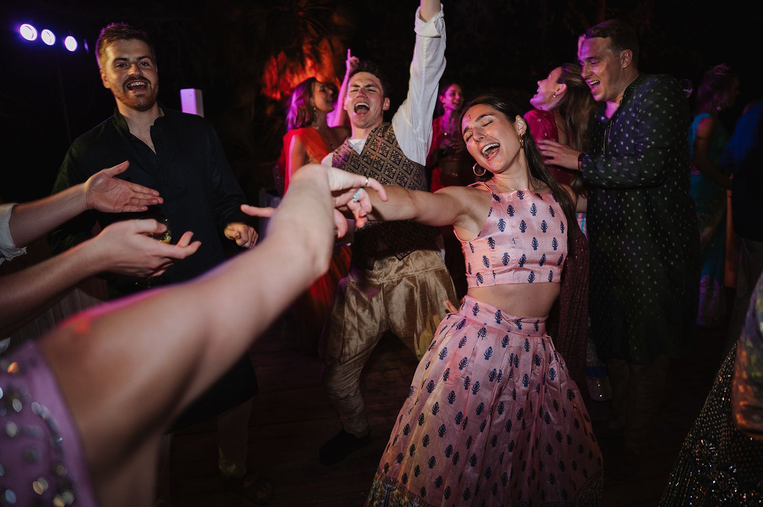 Bride in pink lehenga laughing while dancing with guests at vibrant Indian wedding night reception.