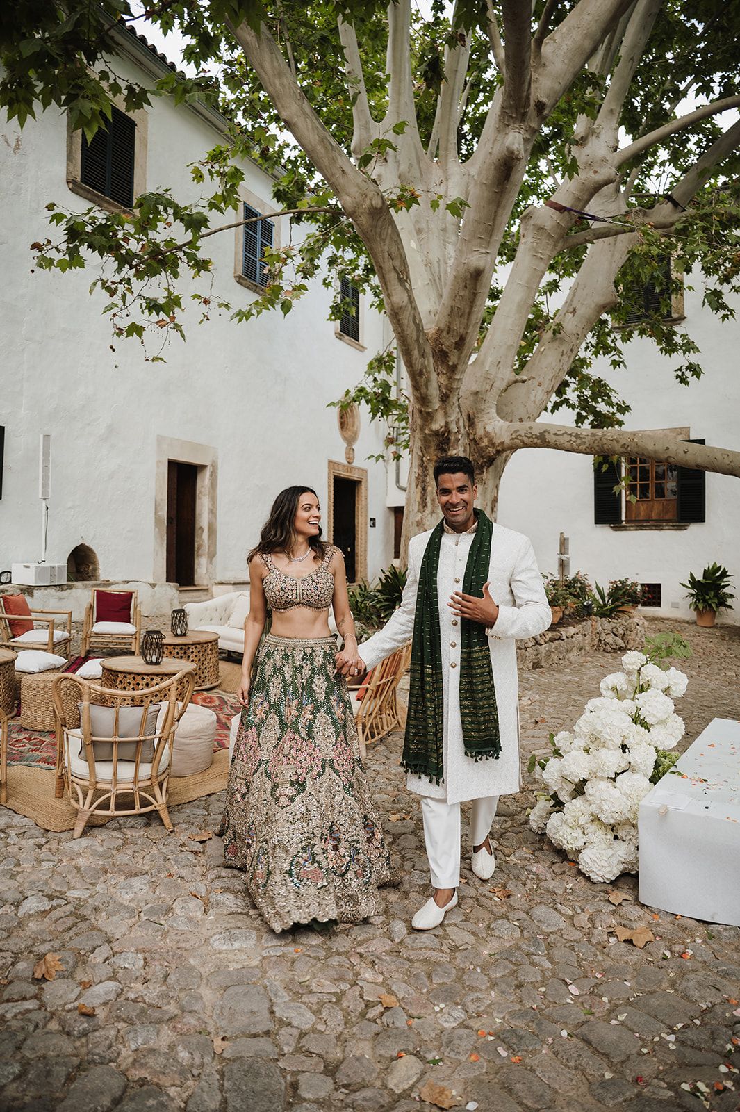 Smiling couple in traditional Indian wedding attire holding hands near a large tree at a villa.