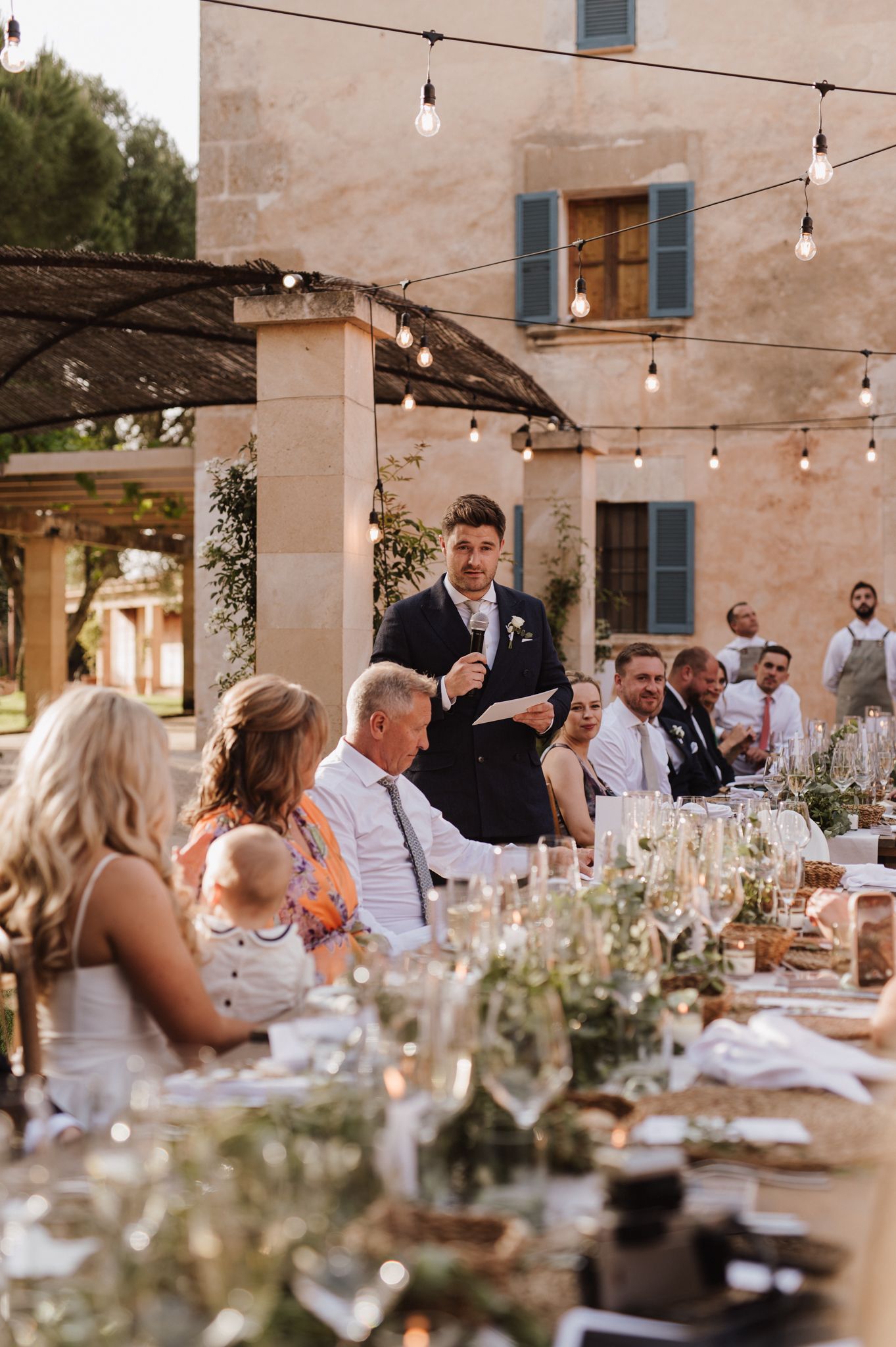 Groom delivering a speech at an outdoor wedding reception under warm string lights.