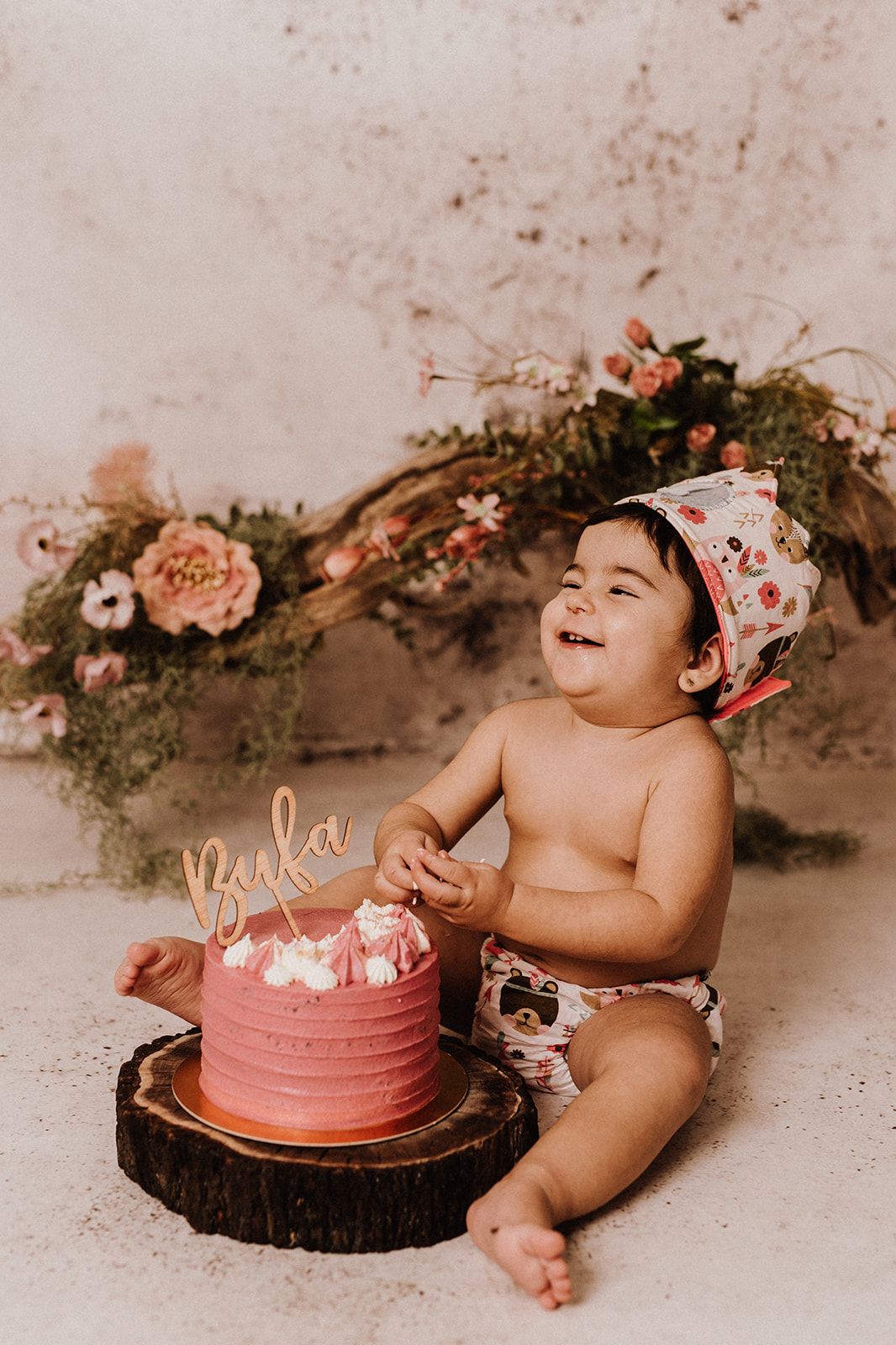 Happy baby girl laughing at her first birthday cake smash with pink frosted cake and floral backdrop.