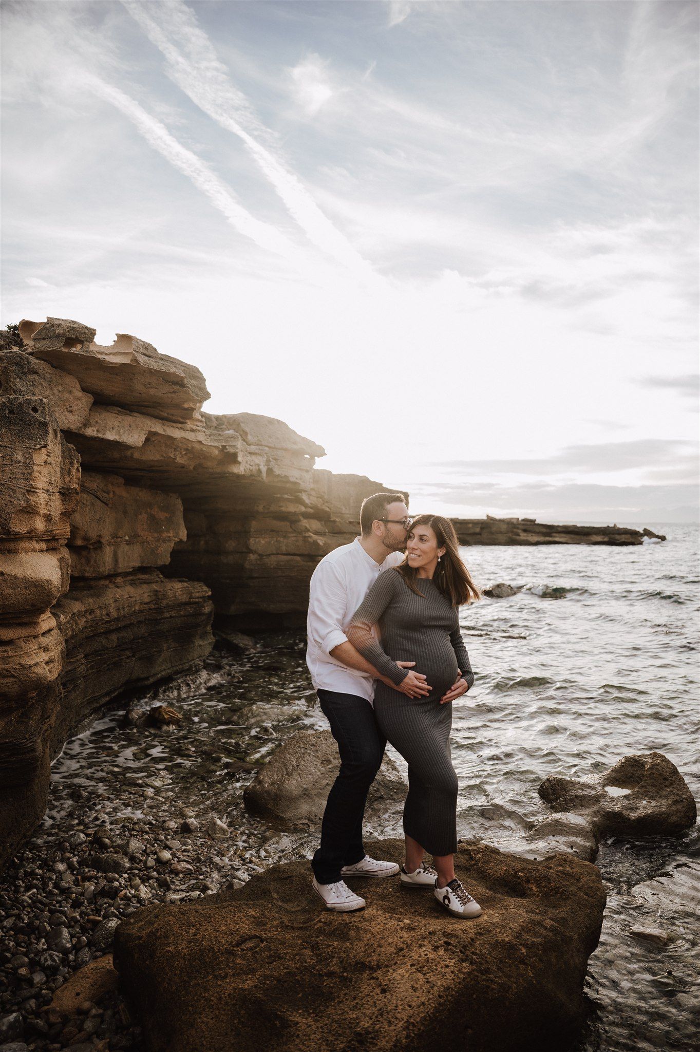 Couple embracing on rocky shoreline at sunset, pregnant woman in gray dress with partner in white shirt.