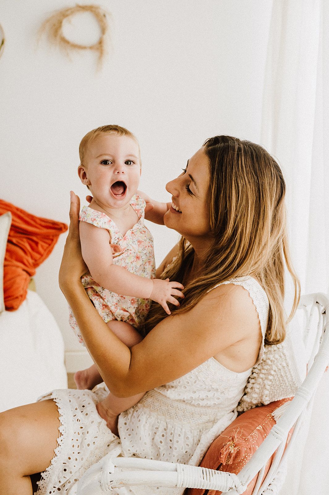 Smiling mother in white dress holds laughing baby girl in floral romper in bright boho bedroom.