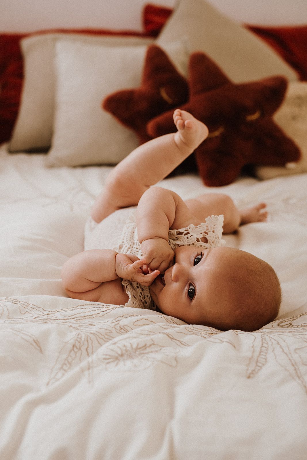 A baby in a lace headband lies on a white bed, surrounded by red and cream pillows, gazing curiously at the camera.