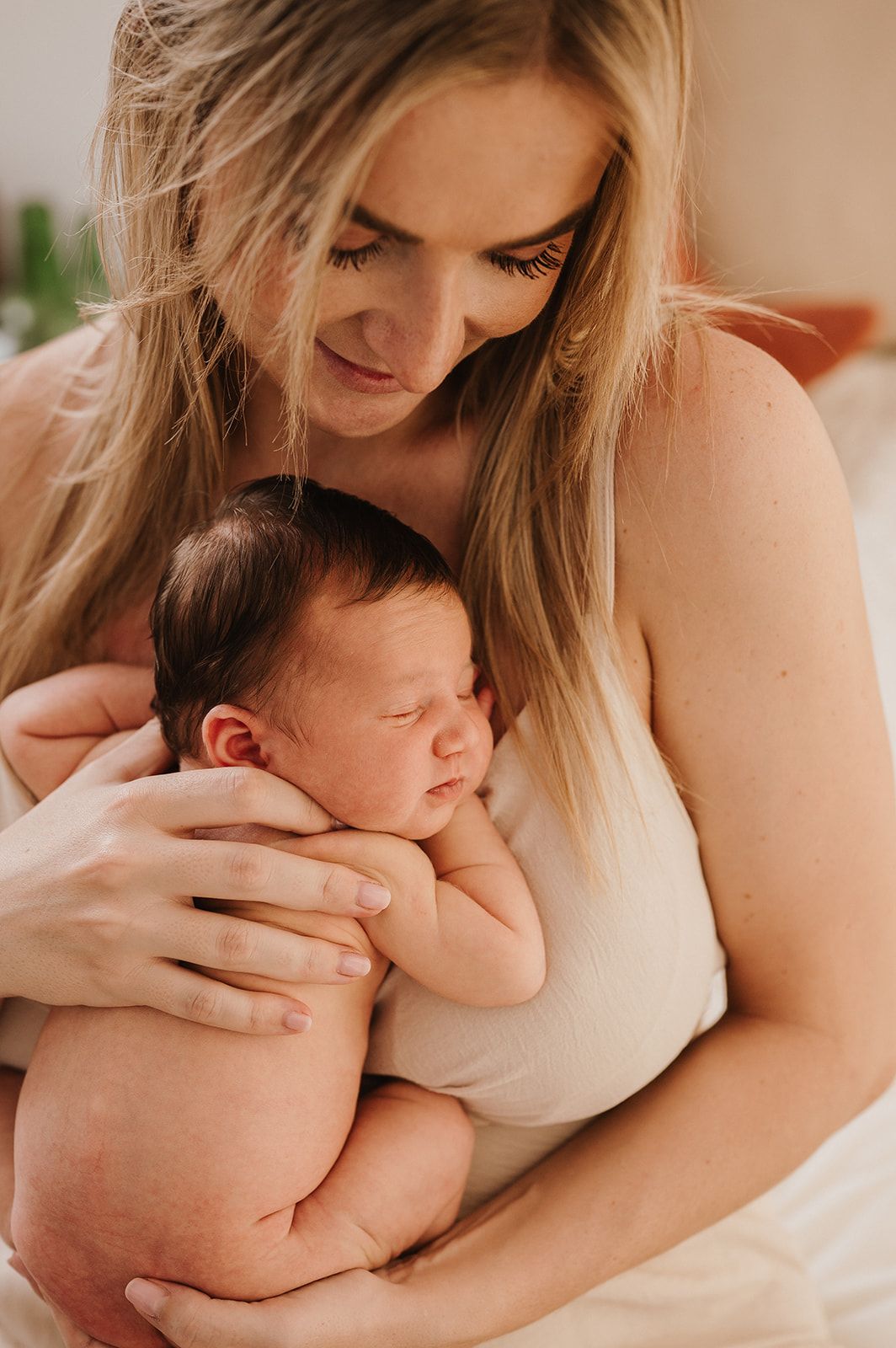 A mother with long blonde hair tenderly cradles her sleeping newborn baby against her chest.