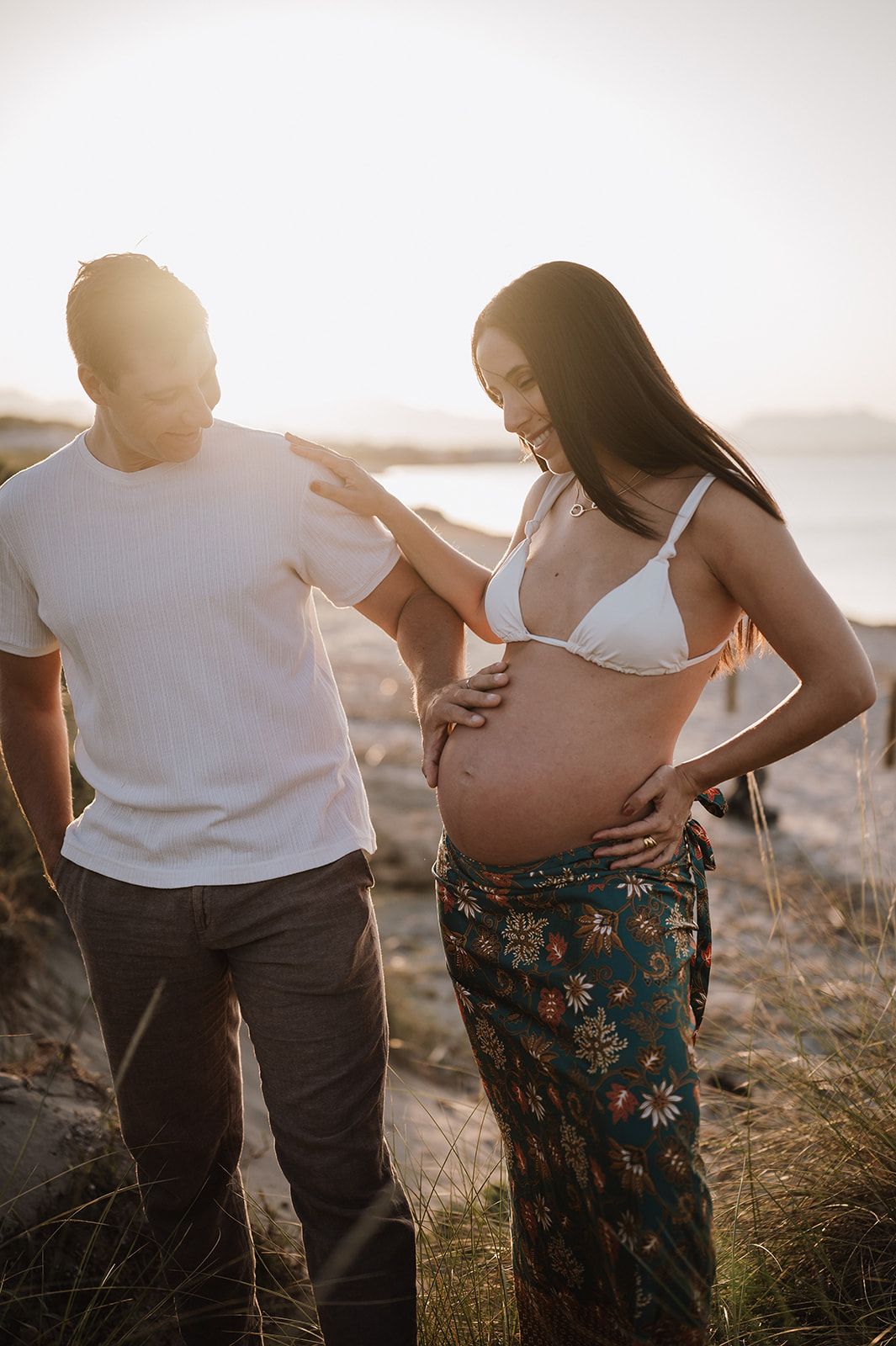 Expectant couple on sunlit beach, man touching pregnant woman's bare bump in floral pants.