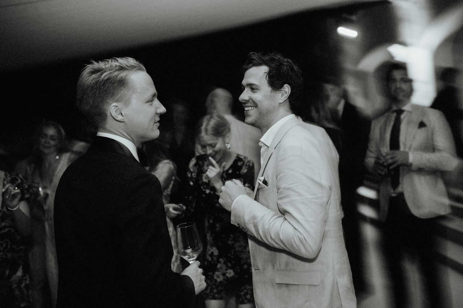 Two men in formal suits laughing together at an elegant evening wedding reception.