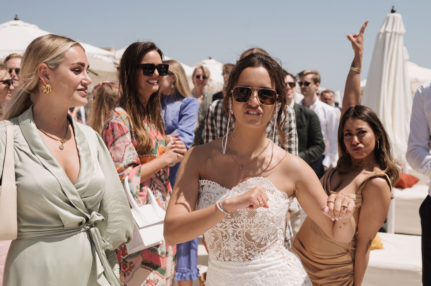 Bride in white lace strapless dress dances joyfully with guests at sunny outdoor wedding reception.