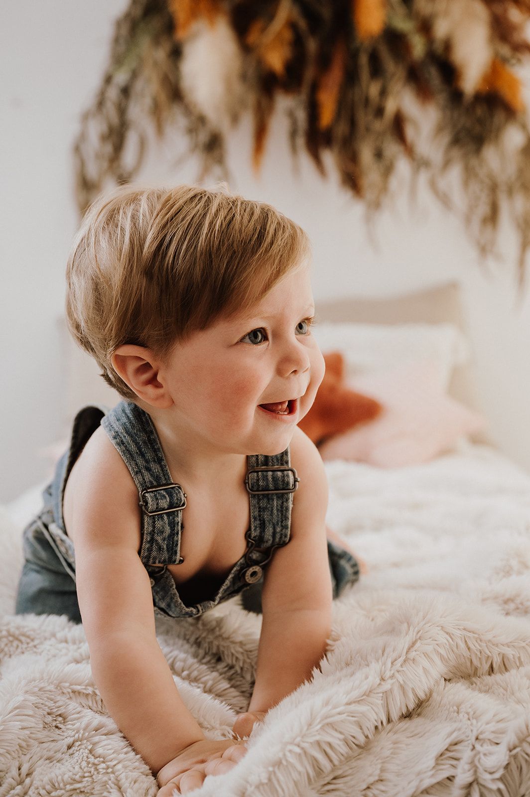 Adorable toddler in denim overalls smiling on a fluffy white blanket with boho floral decor in the background.