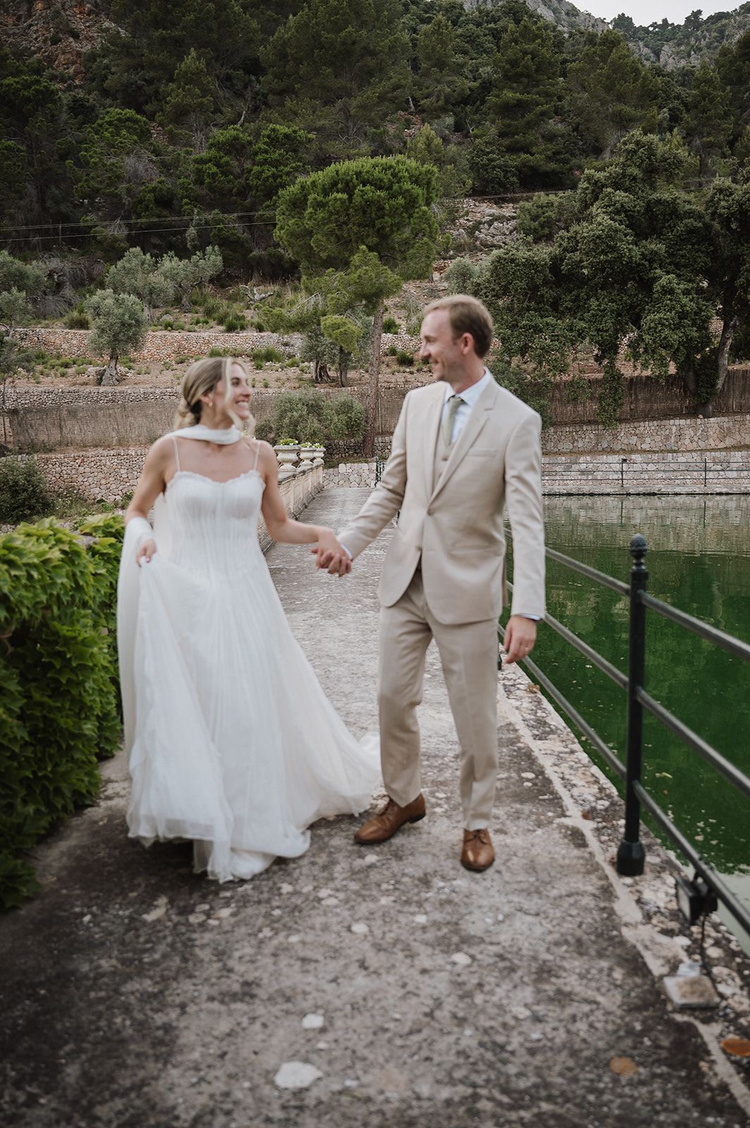 Newlywed couple walks hand in hand beside green water canal at scenic outdoor European wedding venue.