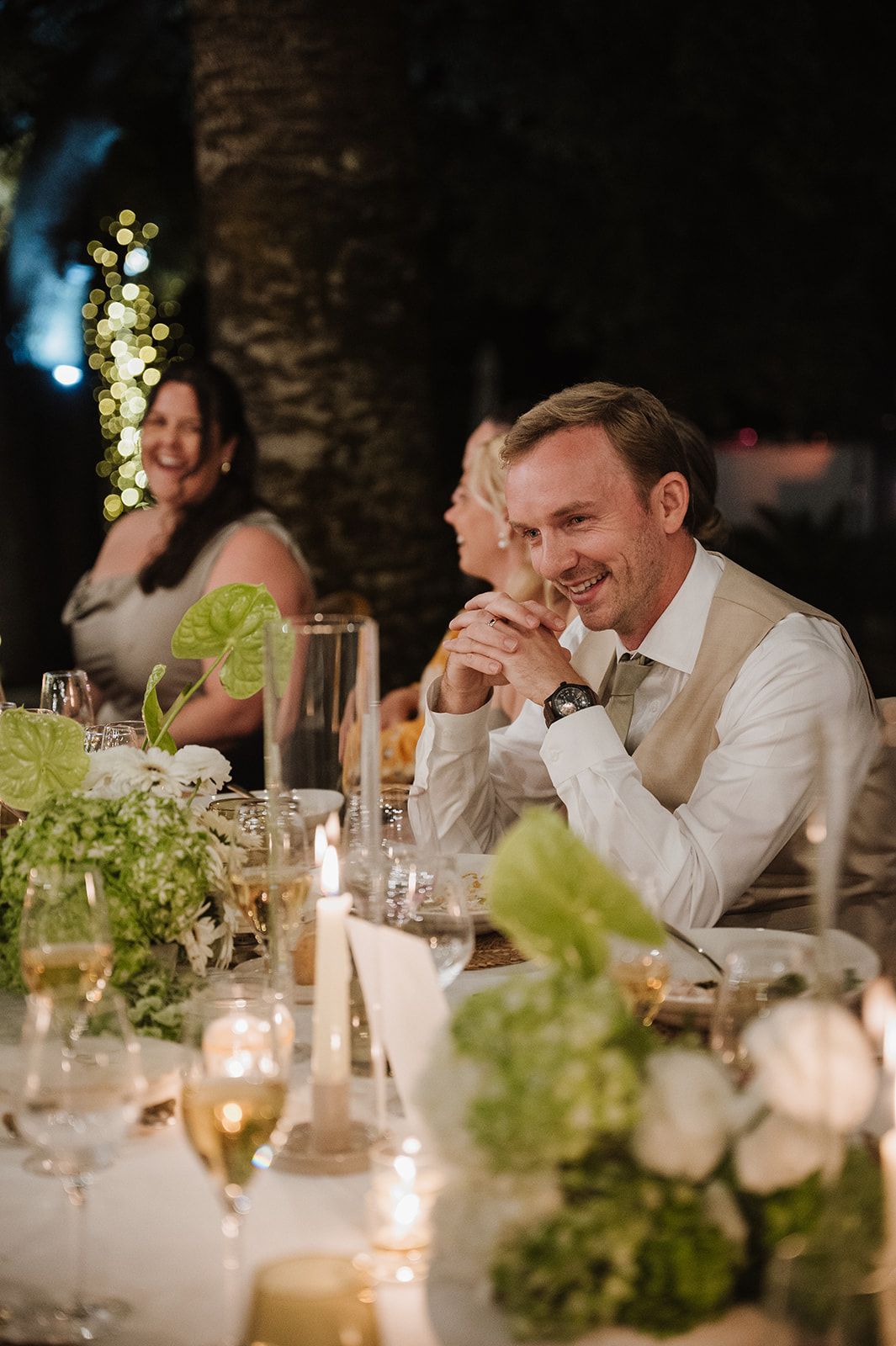 Smiling groom in white shirt and tan vest sits at elegant candlelit wedding reception table with floral centerpieces.