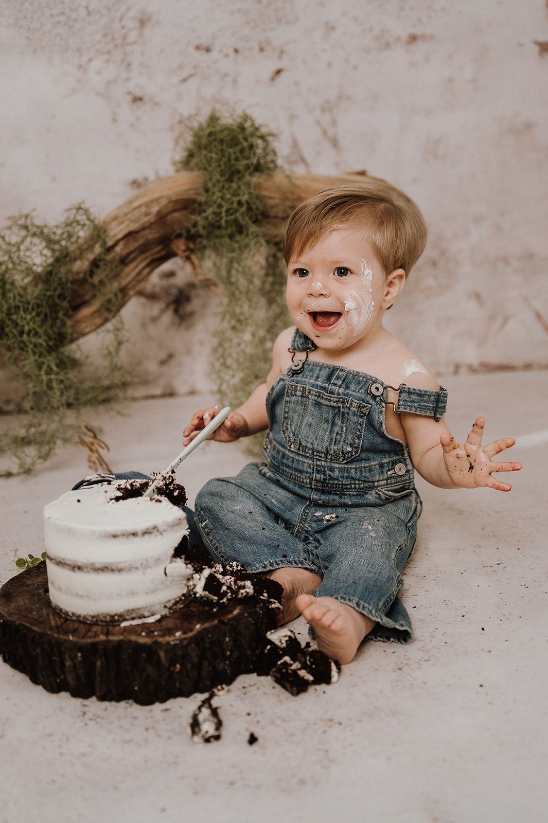 Happy toddler in denim overalls smashing a white cake on a wood slice during a first birthday cake smash photo session.
