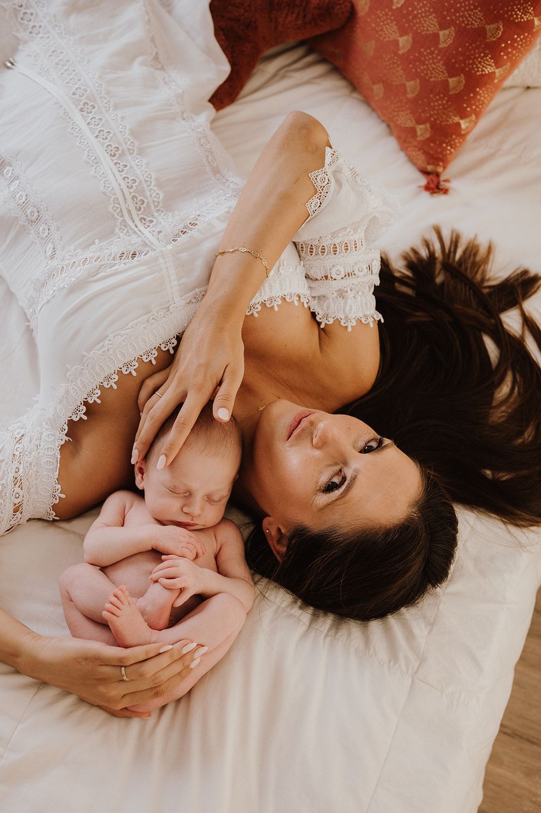 Mother lying on white bed gazing lovingly at her sleeping newborn baby beside her.