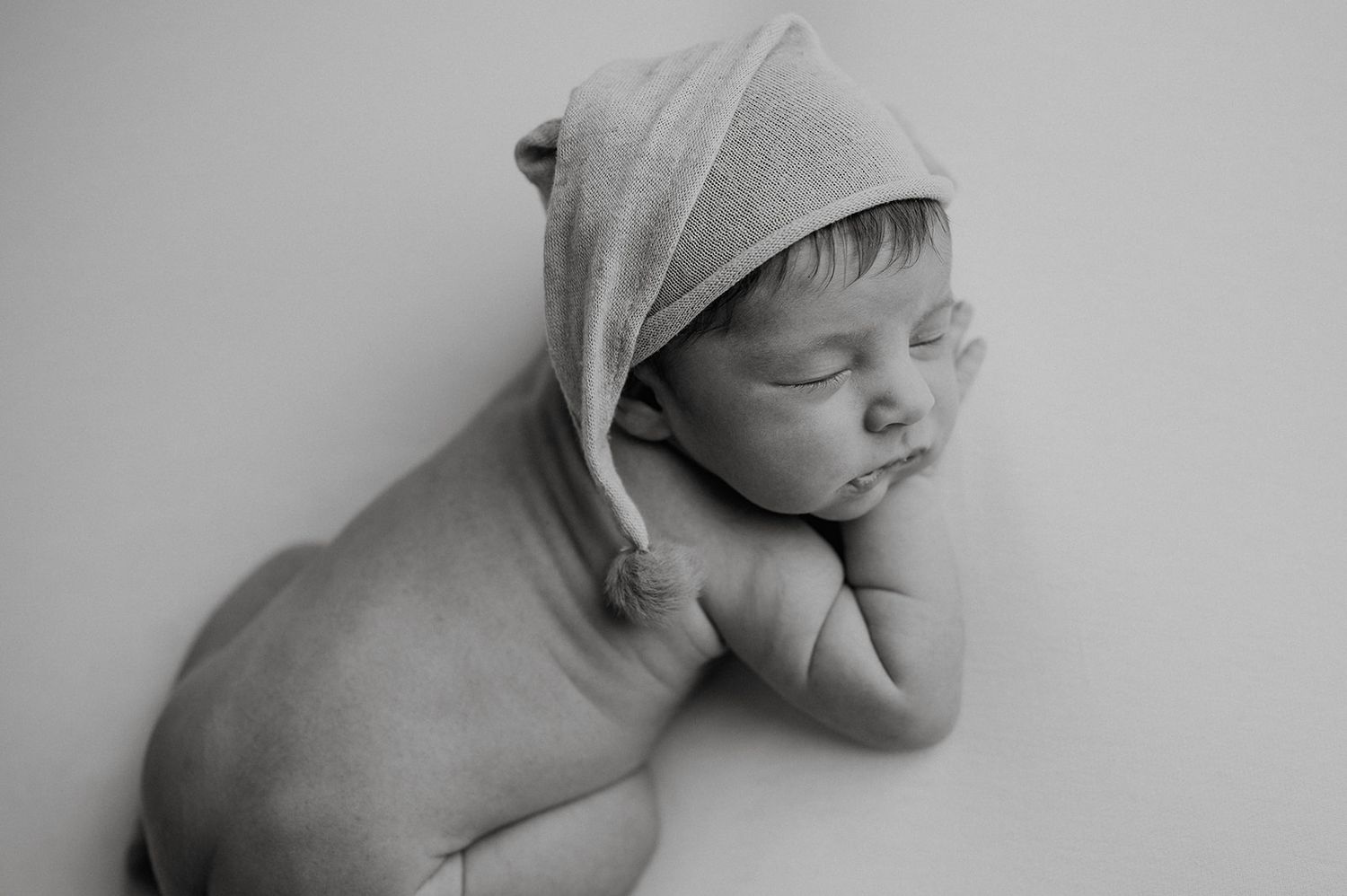 Black and white newborn photo of sleeping baby wearing knit hat, peacefully resting on hands in classic posed portrait.