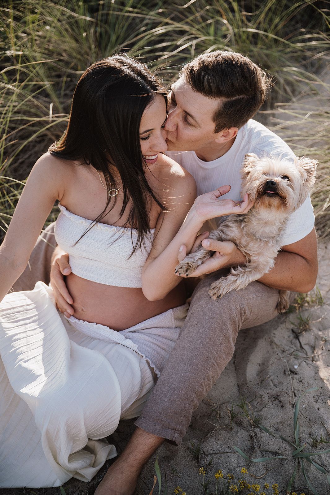 Pregnant couple sitting on sandy beach dunes with small Yorkshire Terrier dog.