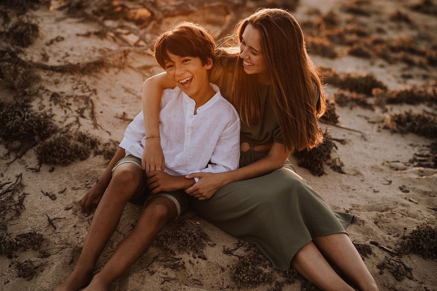 Mother and son laughing together while sitting on sandy beach in warm sunset light.