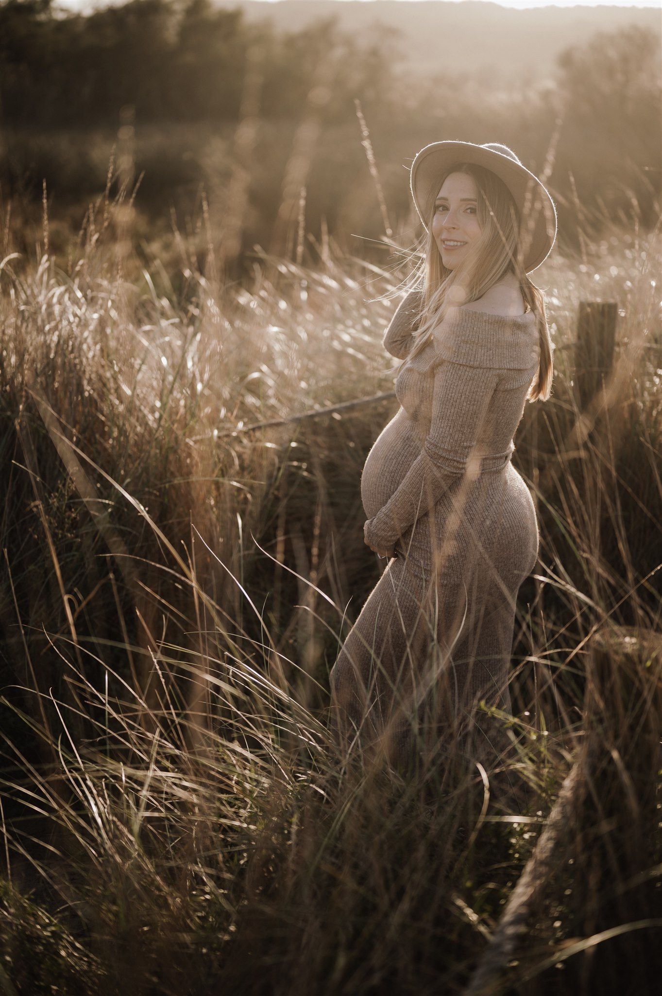 Pregnant woman in gray dress and hat standing in golden field during sunset maternity photoshoot.
