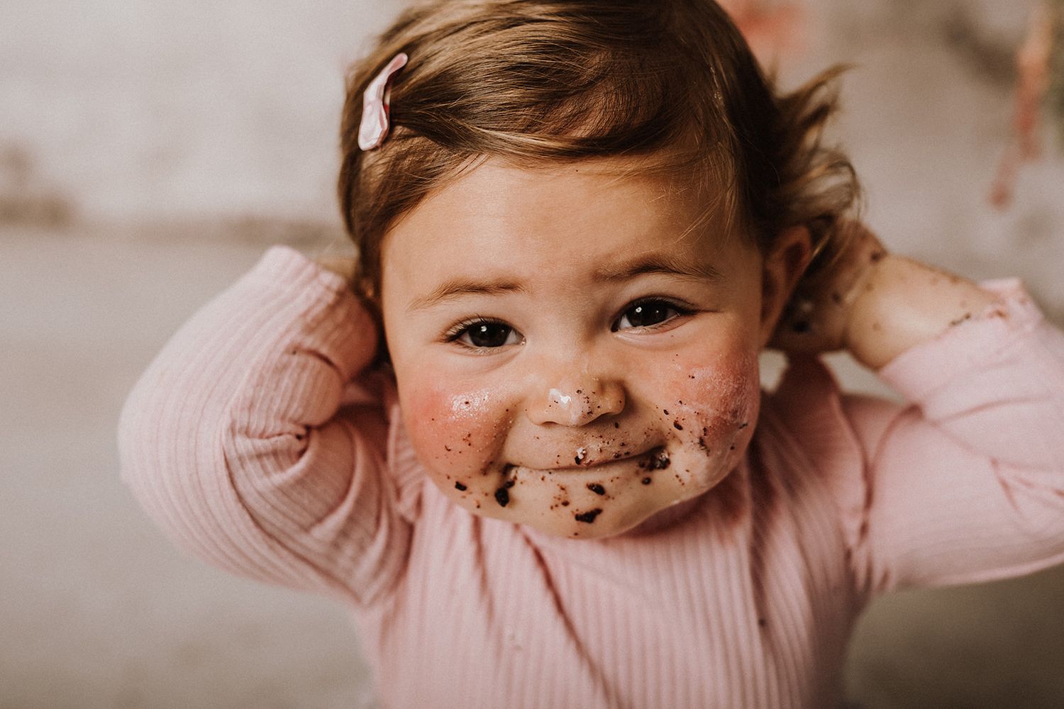 Toddler girl in pink top with chocolate cake smeared on her face, hands raised, smiling during first birthday smash cake session.