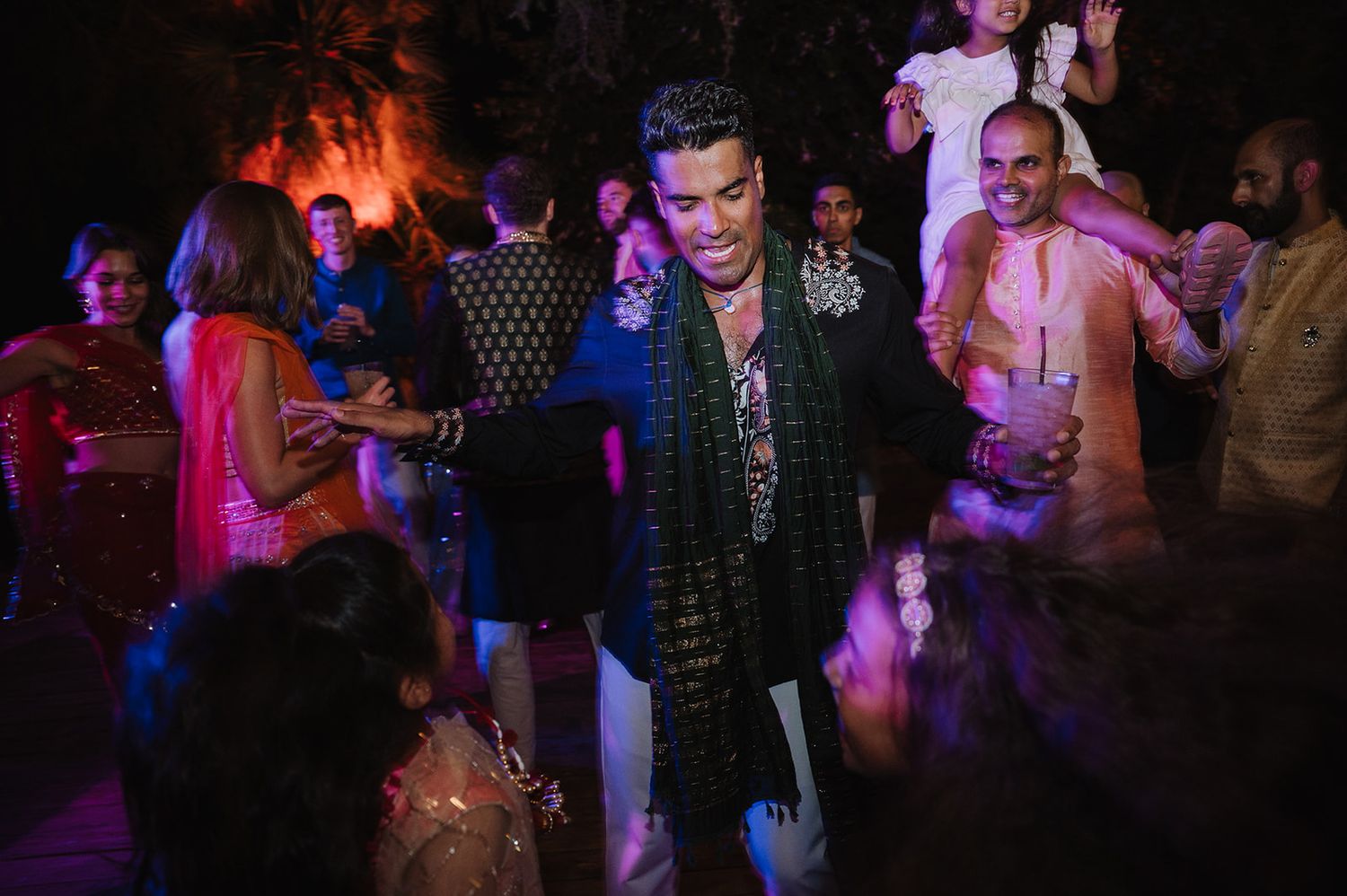 Guests in vibrant Indian attire dance together at a lively wedding reception under colorful party lights.