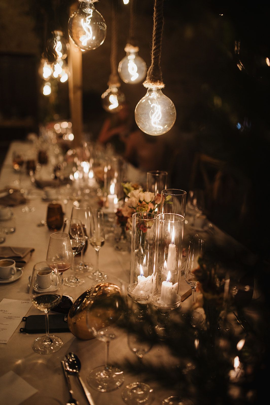 Dimly lit wedding reception table with hanging bulb lights, candles, and crystal glassware.