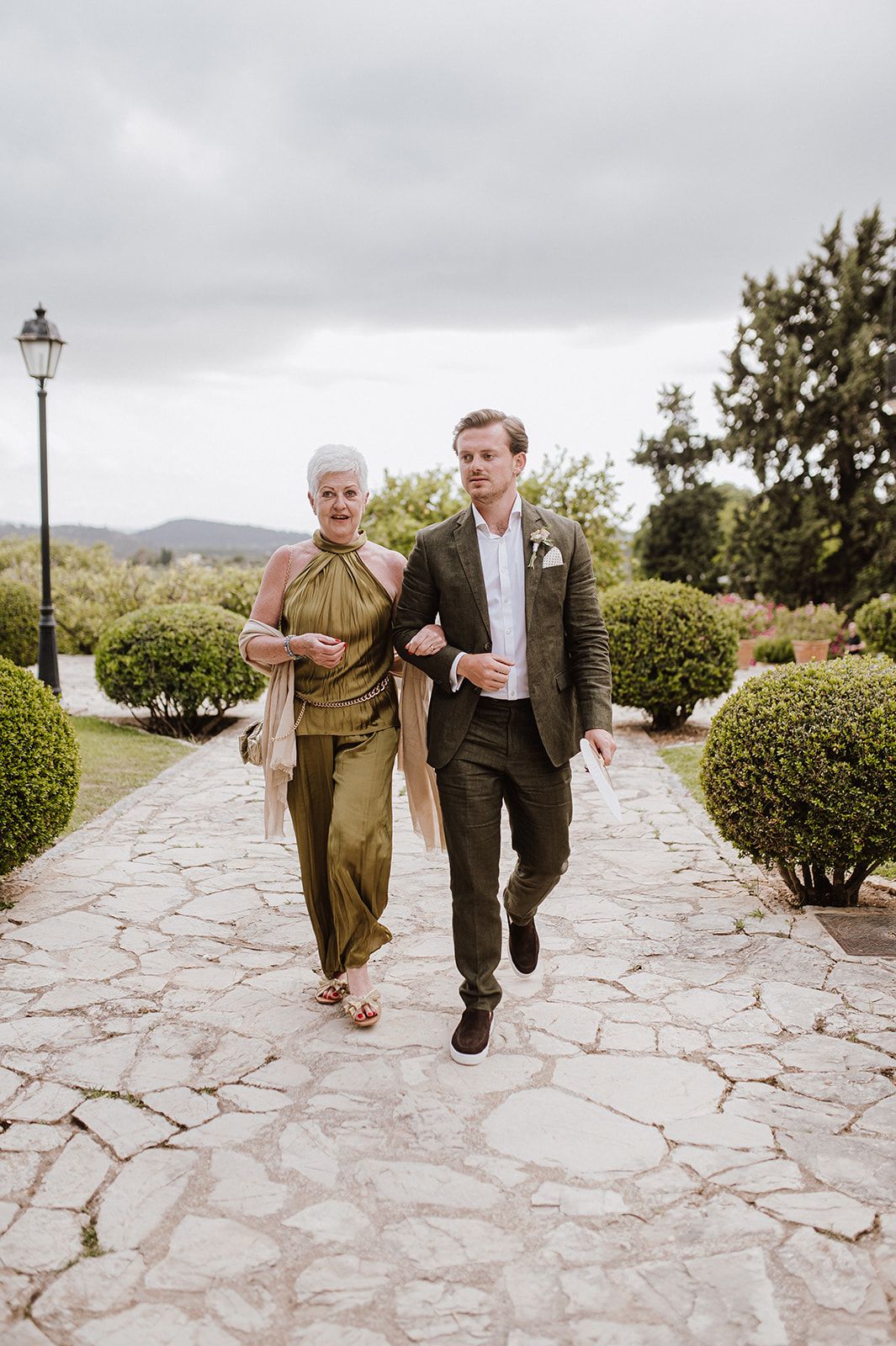 Elegantly dressed couple walk stone garden path, man in brown suit escorts woman in olive jumpsuit.