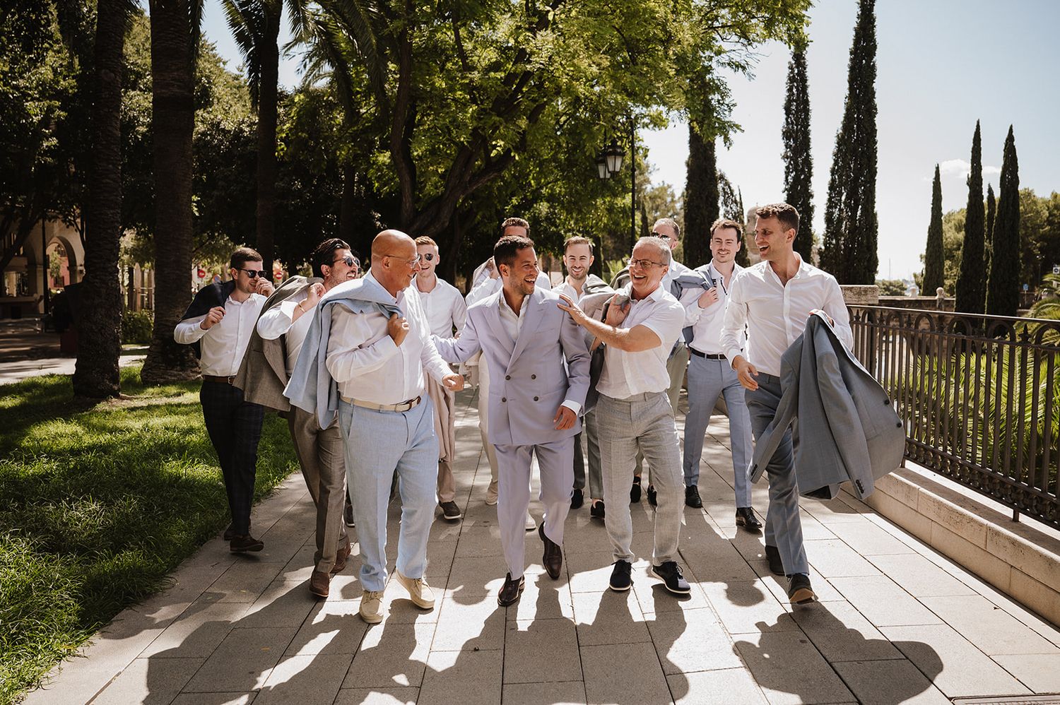Groom and groomsmen in grey suits walking outdoors in sunlit garden before wedding.