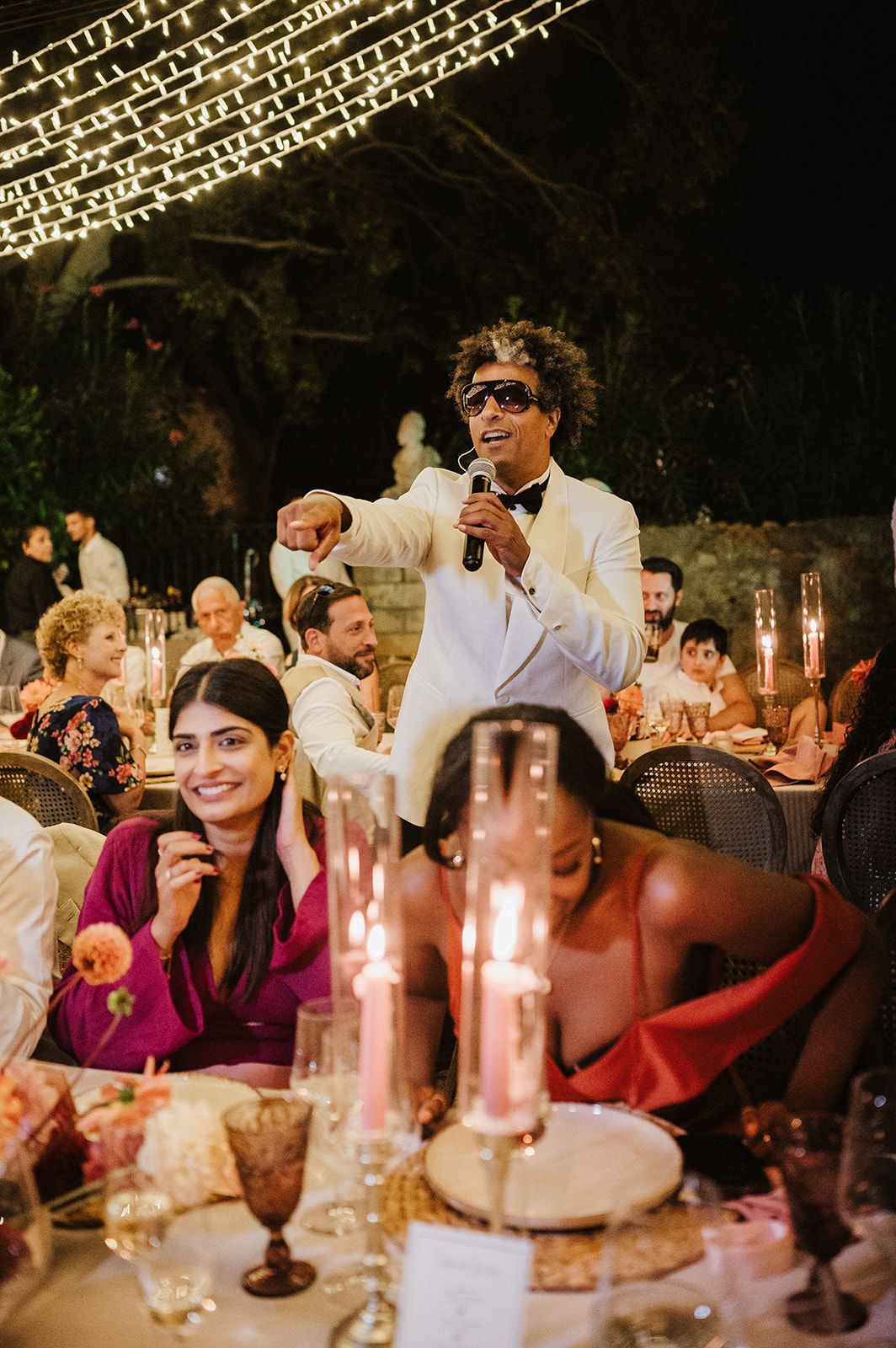 Energetic wedding entertainer in white tuxedo performs with microphone at candlelit evening reception.