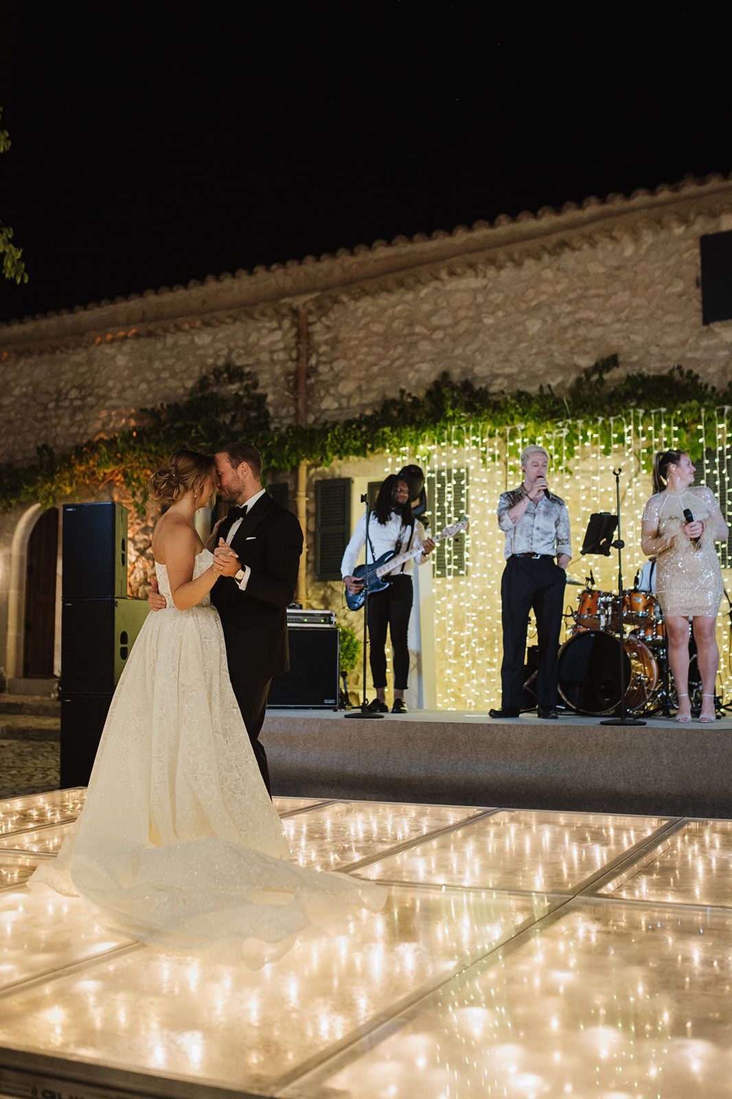Bride and groom share first dance on illuminated floor while live band performs at night wedding.
