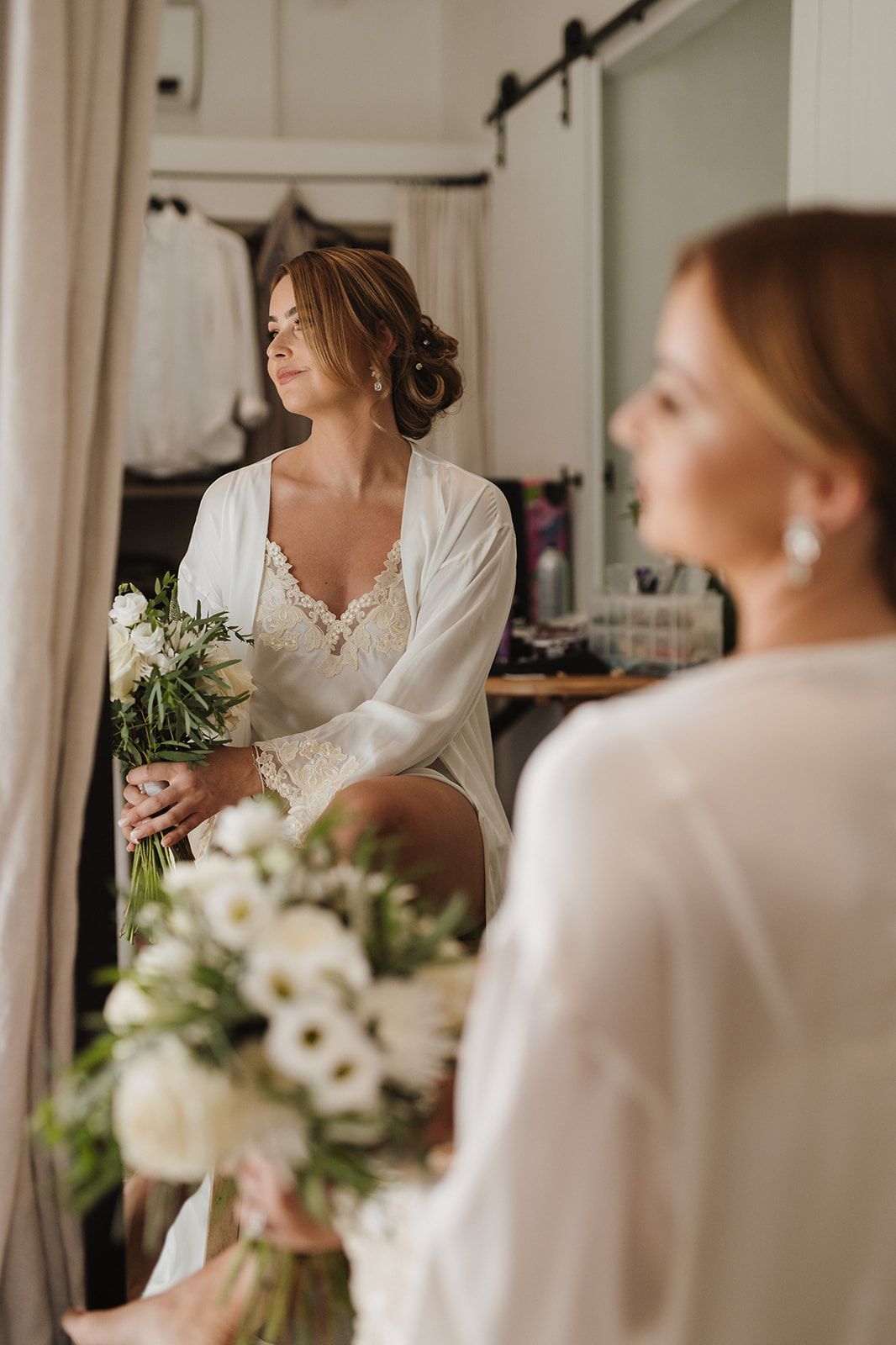 Elegant bride in white robe holds a bouquet of white flowers while gazing into a mirror.