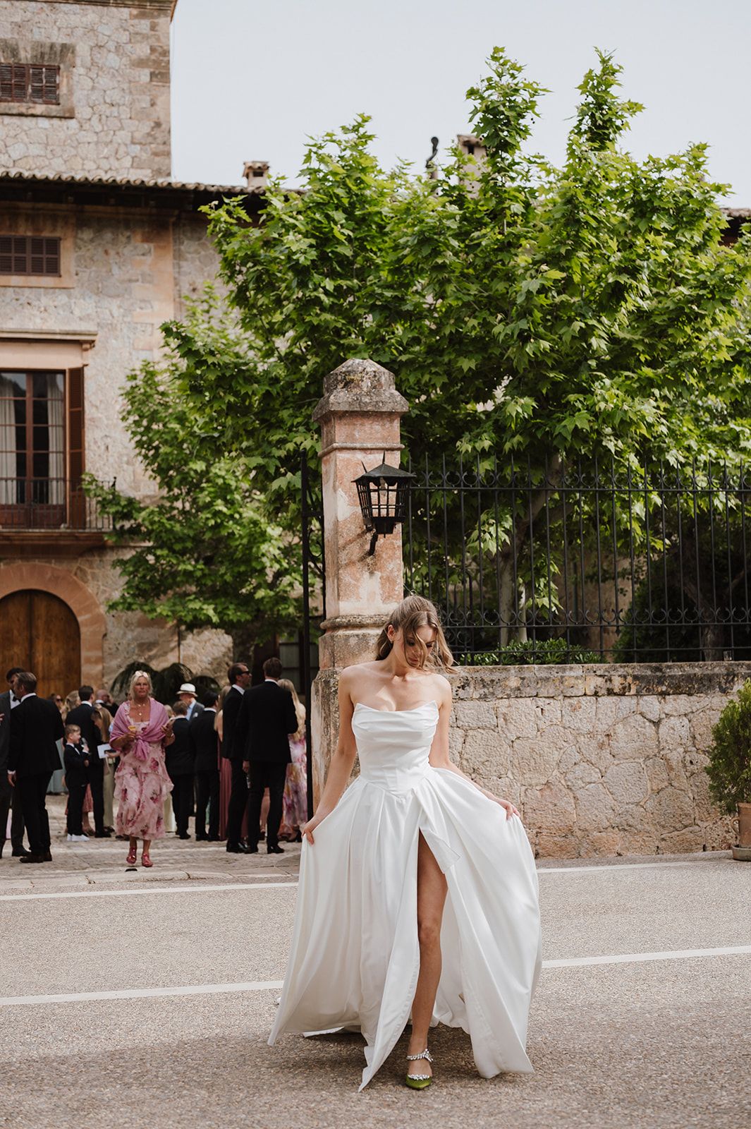 Bride in strapless white gown with high slit walks outside rustic stone venue with wedding guests in background.