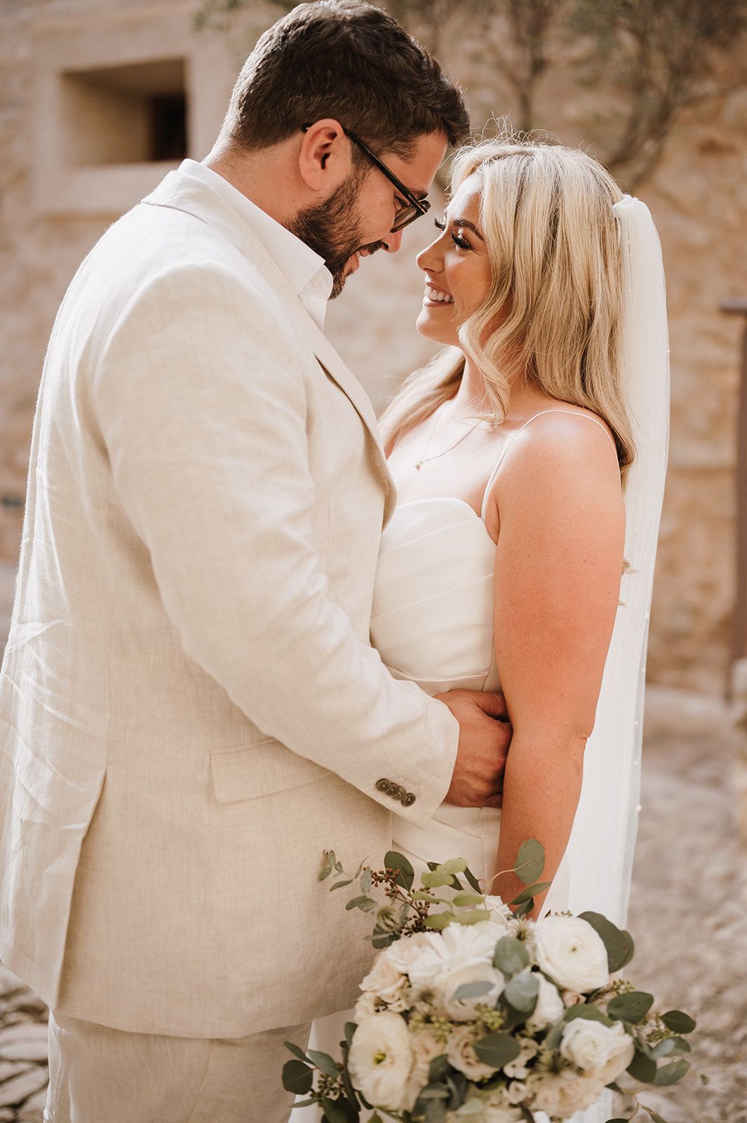 Bride and groom embracing closely holding white floral bouquet at rustic stone venue.