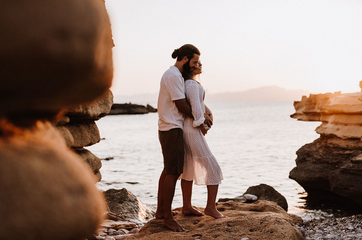 Couple embracing on rocky beach at golden sunset with calm ocean and mountains in background.