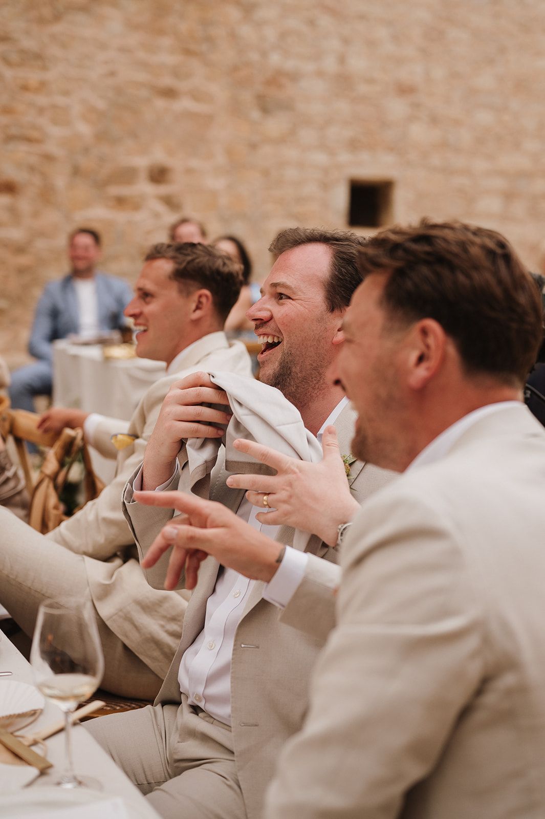 Wedding guests in beige suits laughing joyfully during speeches at an elegant reception dinner.
