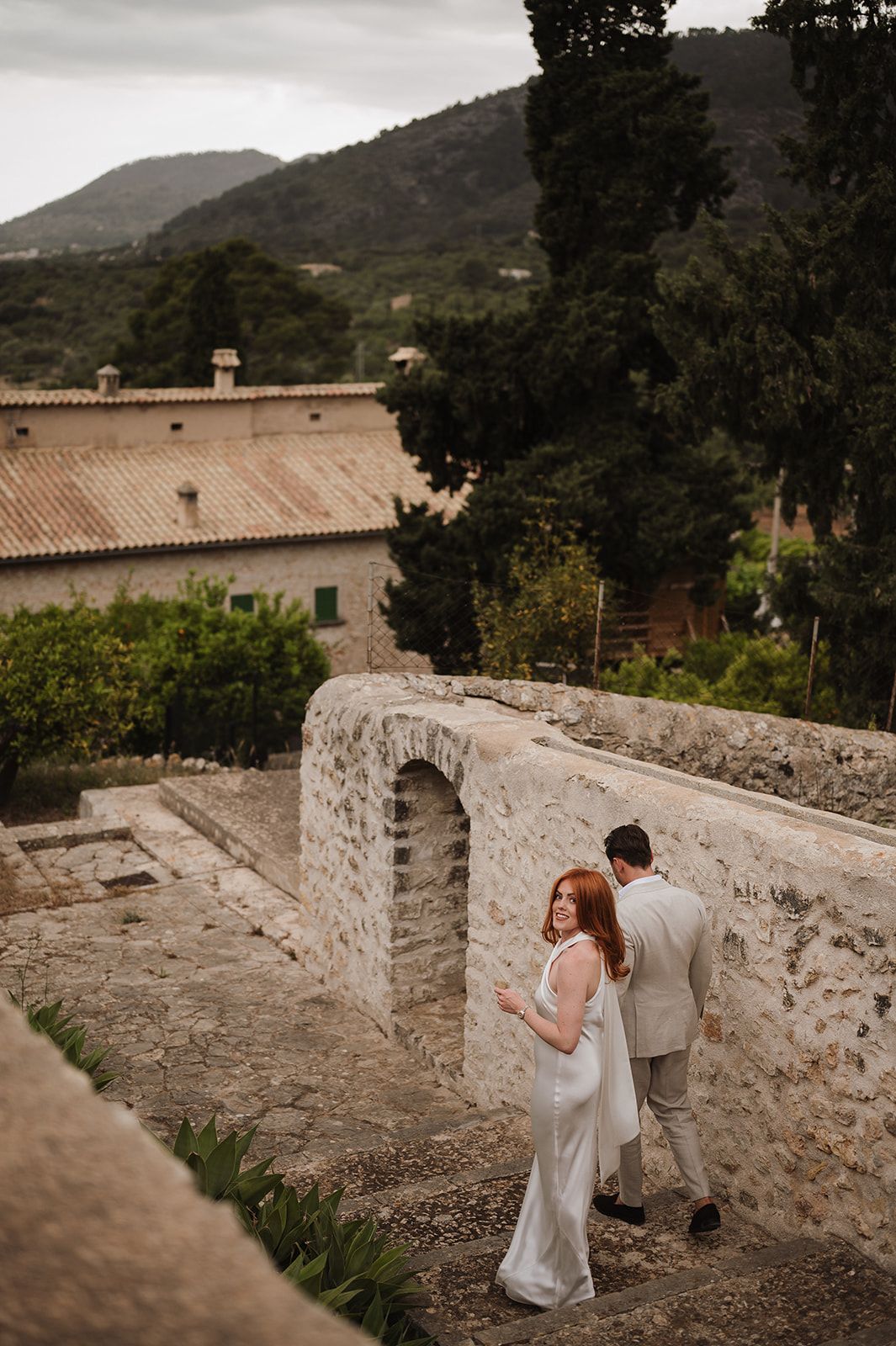 Bride and groom explore rustic stone architecture with lush green hillside village in the background.