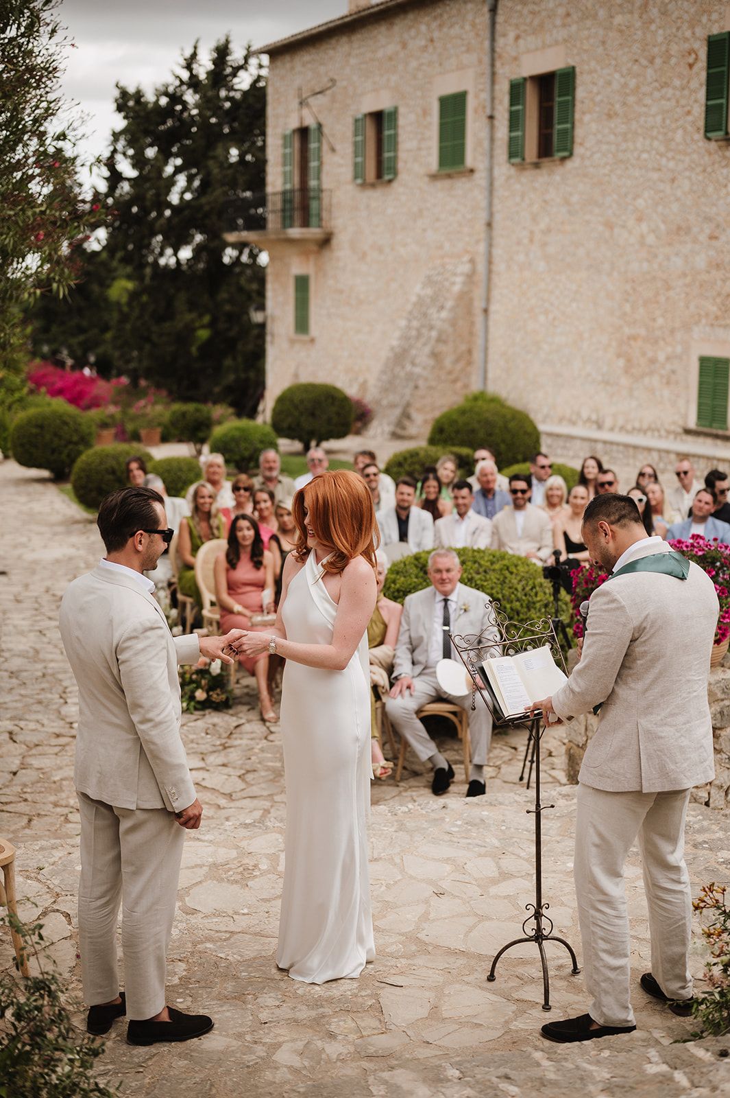Couple exchanges vows at outdoor wedding ceremony with stone villa, manicured hedges, and seated guests.