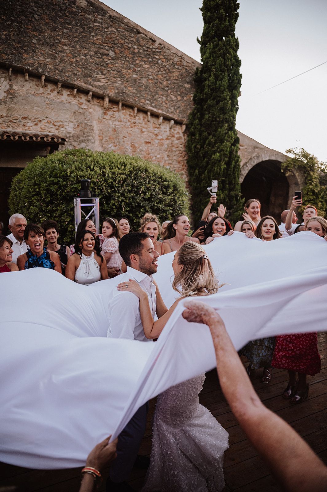 Bride and groom kiss under billowing white sheet surrounded by cheering wedding guests.