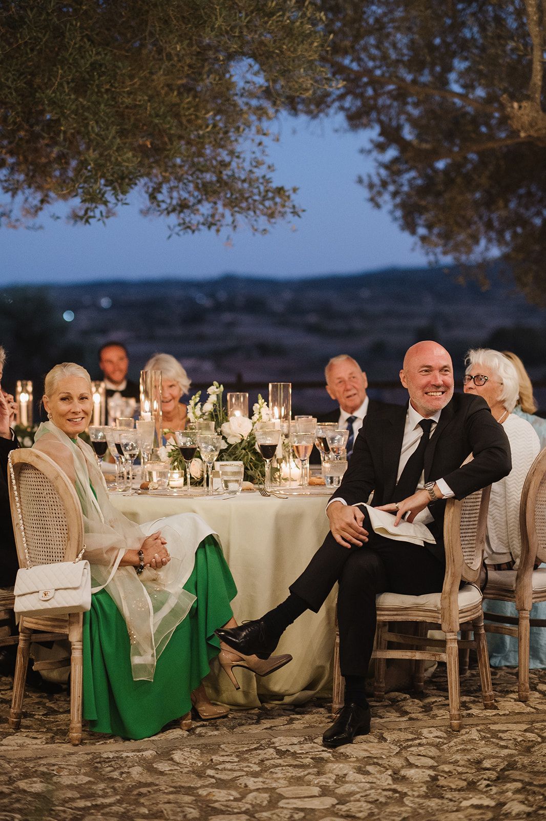 Wedding guests smile at candlelit outdoor reception table at dusk with scenic hillside views in background.