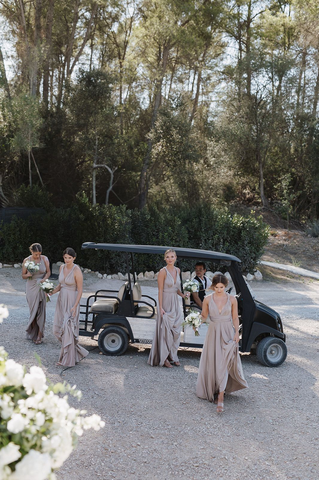 Bridesmaids in taupe wrap dresses walking past golf cart on gravel path at wedding.
