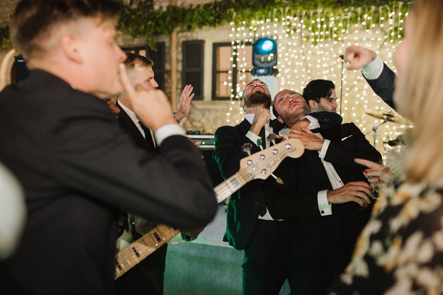 Energetic wedding band performers in tuxedos play electric guitar enthusiastically on stage at evening reception.