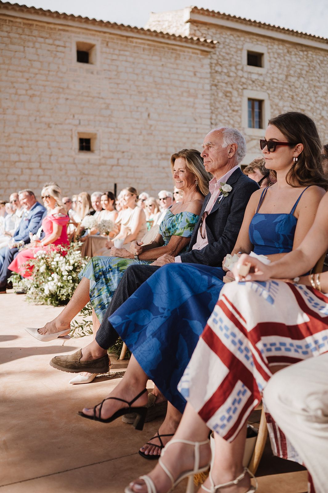 Wedding guests seated outdoors at a rustic stone venue ceremony on a sunny day in colorful summer attire.