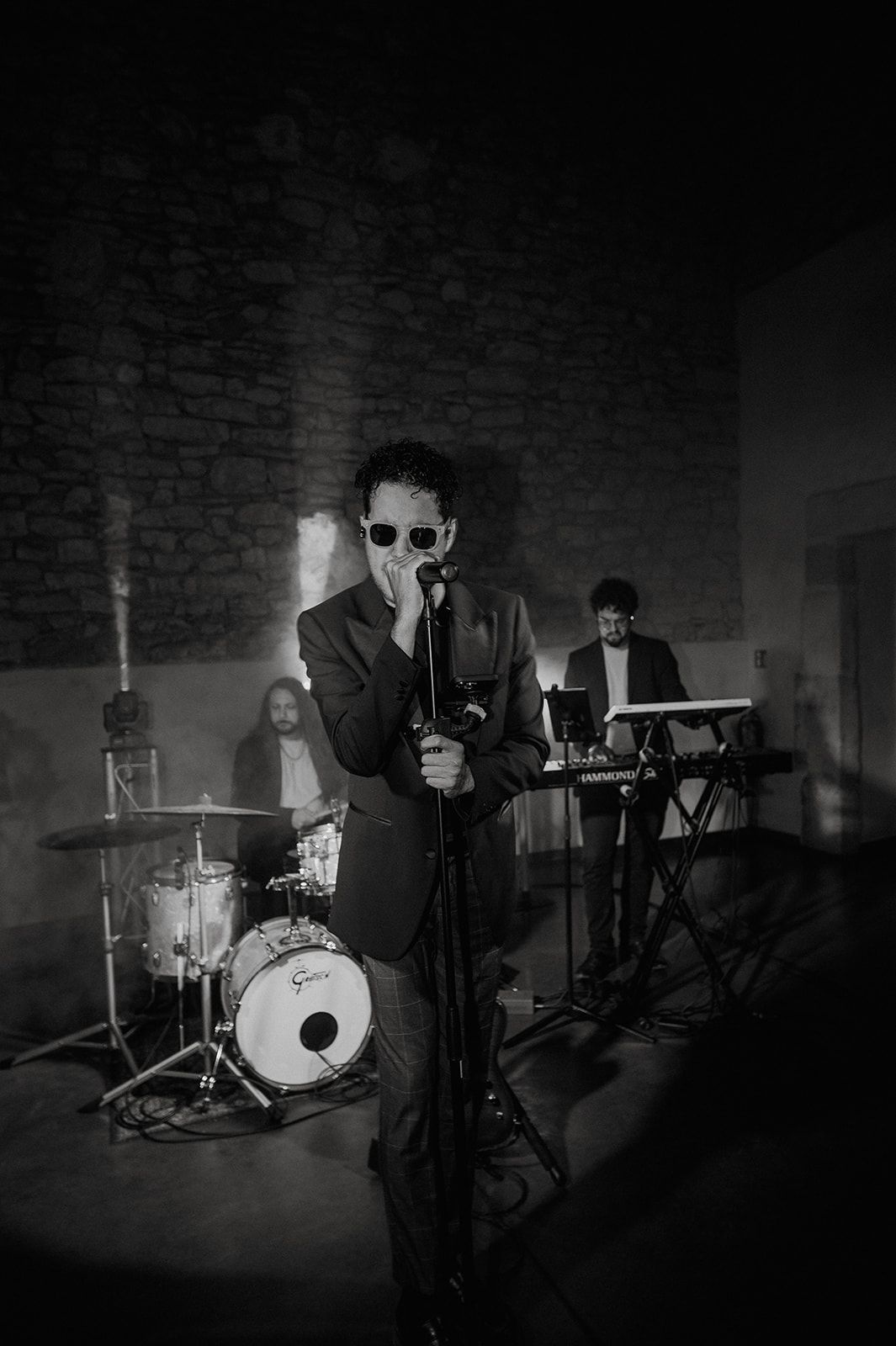 Black and white photo of a band performing live, with a suited vocalist at mic, drummer, and keyboardist in a stone venue.